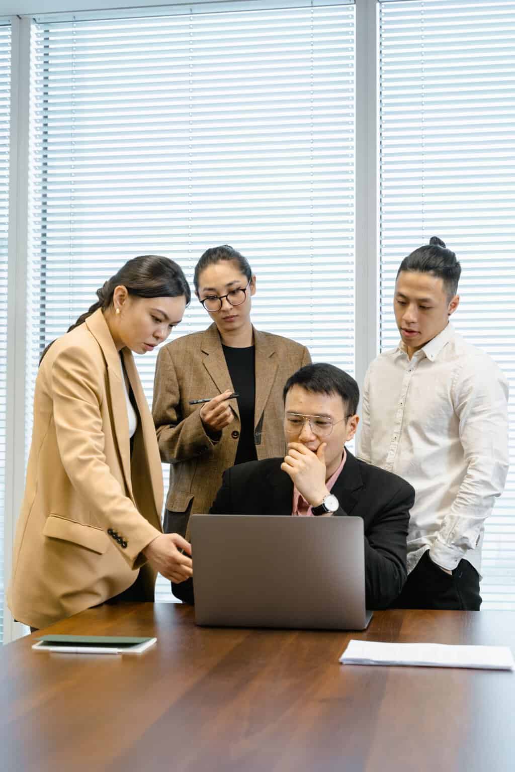 A group of colleagues engaged in a meeting, discussing around a laptop in a modern office.